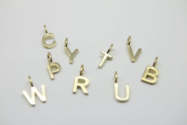 May include: A set of gold-colored metal letter charms, including the letters B, C, P, R, T, U, V, W, and Y. The charms are all in a simple, modern font.