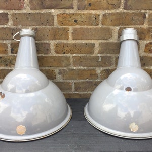 May include: Two vintage industrial pendant lights with a grey enamel finish. The lights have a dome shape with a narrow neck and a metal ring at the top.
