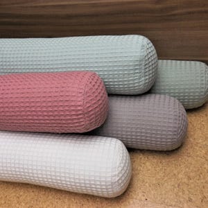 Könnte beinhalten: Vier zylindrische Kissen in verschiedenen Farben: hellblau, rosa, grau und weiß. Die Kissen haben eine Waffelstruktur und sind übereinander gestapelt.
