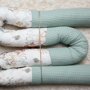 Könnte beinhalten: Ein dreiteiliges Baby-Kissen-Set. Das Kissen ist aus hellgrünem Waffelstoff mit einem gemusterten Stoff mit Hirschen und Waldelementen. Das Set ist mit Bindfaden zusammengebunden. Das Kissen ist etwa 152 cm lang.