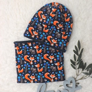 Könnte beinhalten: Ein marineblaues Mützen- und Halstuch-Set mit einem sich wiederholenden Muster aus orangefarbenen Füchsen, braunen Eichhörnchen, Bäumen und blauen Akzenten. Das Set ist aus weichem Stoff gefertigt.
