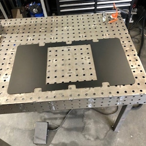 May include: A black metal plate with a cut-out design, sitting on a metal work table with a grid pattern. The plate is used for welding or fabrication.