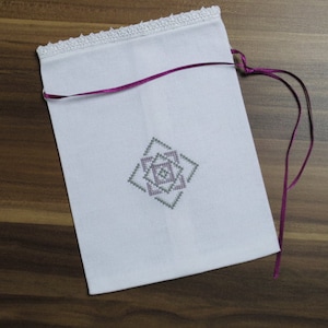 Könnte beinhalten: Ein weißer Stoffbeutel mit einem Kreuzstich-Geometriedesign in Grau- und Lavendeltönen. Der obere Rand hat eine weiße Spitzentrimmung, und ein lila Band ist zum Verschließen eingefädelt. Der Beutel liegt auf einer Holzoberfläche.