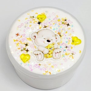 May include: White slime with yellow and pink glitter and a white bear holding a yellow bear. The slime is in a clear plastic container.