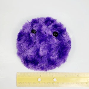 K&ouml;nnte beinhalten: Ein lila Pl&uuml;schtier in Form einer Kugel mit zwei schwarzen Augen. Das Spielzeug hat einen Durchmesser von etwa 7,5 cm.