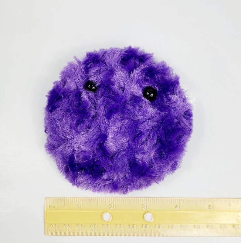 K&ouml;nnte beinhalten: Ein lila Pl&uuml;schtier in Form einer Kugel mit zwei schwarzen Augen. Das Spielzeug hat einen Durchmesser von etwa 7,5 cm.