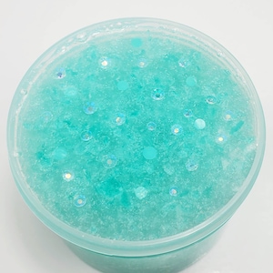 May include: A close-up of a light blue slime with iridescent glitter and white flecks. The slime is in a clear plastic container.