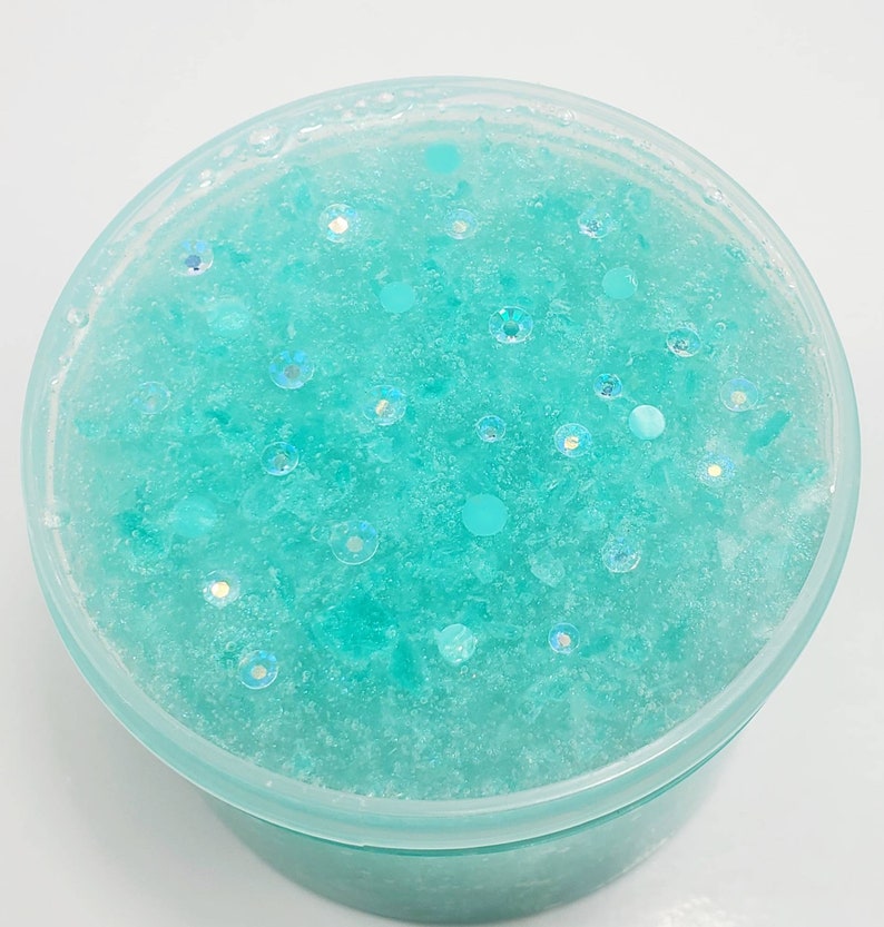 May include: A close-up of a light blue slime with iridescent glitter and white flecks. The slime is in a clear plastic container.