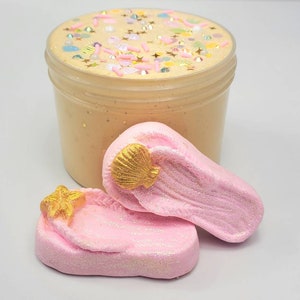 May include: A clear plastic jar filled with light peach-colored slime topped with colorful sprinkles, small gold stars, and iridescent beads, accompanied by two pink flip-flop shaped bath bombs featuring gold-colored seashell and starfish accents.