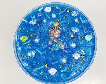 May include: Blue slime with glitter and various mermaid-themed decorations, including a mermaid figurine, seashells, and pearls.