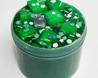 May include: Green slime with green and white sprinkles, green and white star-shaped sprinkles, and a small frog figurine. The slime is in a clear plastic container.