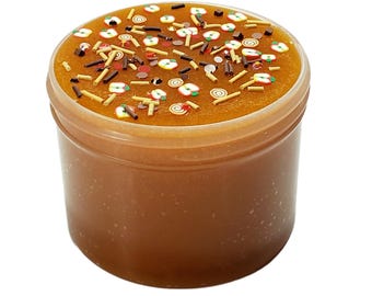 May include: A clear plastic container filled with brown slime, topped with decorative sprinkles. The sprinkles include apple shapes, brown sticks, and small red and white pieces. The slime has a glossy, smooth texture.