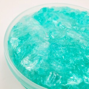 May include: A close-up of a teal blue slime with a glittery finish in a clear plastic container.