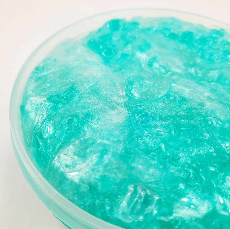 May include: A close-up of a teal blue slime with a glittery finish in a clear plastic container.
