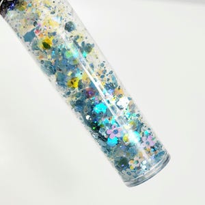 May include: A clear, cylindrical container filled with a mix of colorful glitter. The glitter includes shades of blue, yellow, and iridescent pieces. The container is angled, showcasing the glitter's sparkle and the clear material of the container.