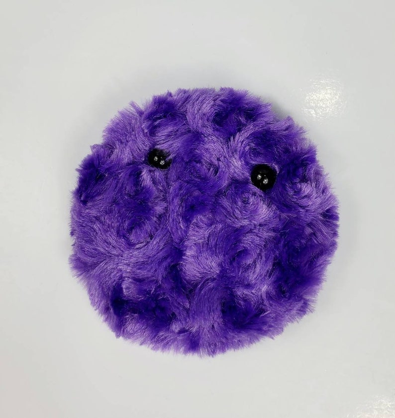 K&ouml;nnte beinhalten: Ein rundes Pl&uuml;schtier in Lila mit zwei schwarzen Augen. Das Spielzeug ist aus weichem, flauschigem Material.