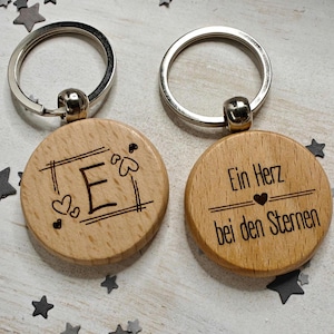 Könnte beinhalten: Zwei Holzschlüsselanhänger mit silbernen Schlüsselringen. Ein Schlüsselanhänger hat den Buchstaben "E" in einem quadratischen Rahmen mit Herzen graviert. Der andere Schlüsselanhänger hat den Text "Ein Herz bei den Sternen" mit einem kleinen Herzen graviert.