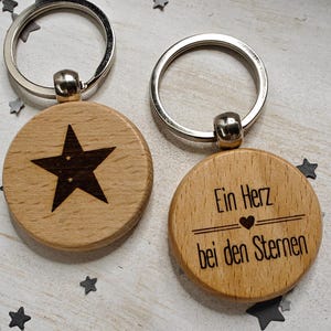 Könnte beinhalten: Zwei Holzschlüsselanhänger mit Metallringen. Ein Schlüsselanhänger hat einen schwarzen Stern und der andere den Text "Ein Herz bei den Sternen" in Schwarz.