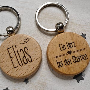 Könnte beinhalten: Zwei Holzschlüsselanhänger mit silbernen Schlüsselringen. Ein Schlüsselanhänger hat den Namen "Elias" mit einem kleinen Herzsymbol eingraviert. Der andere Schlüsselanhänger hat den Text "Ein Herz bei den Sternen" mit einem kleinen Herzsymbol eingraviert.