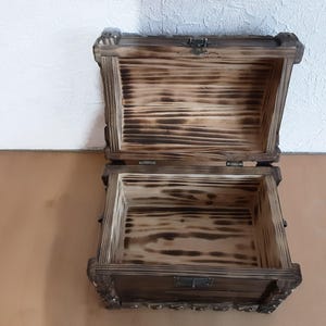May include: A wooden treasure chest with a hinged lid and a metal latch. The chest is made of dark wood with a natural wood grain and has a distressed finish. The inside of the chest is lined with a light wood.