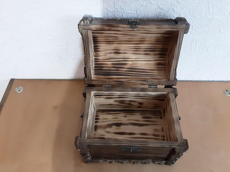 May include: A wooden treasure chest with a hinged lid and a metal latch. The chest is made of dark wood with a natural wood grain and has a distressed finish. The inside of the chest is lined with a light wood.