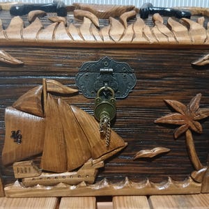 May include: Wooden treasure chest with a nautical theme. The chest is carved with a sailboat, palm tree, and waves. It has a metal clasp and a lock with a key. The chest is brown with a natural wood grain.