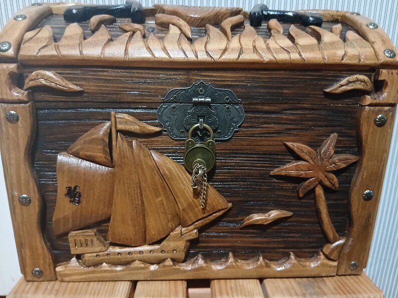 May include: Wooden treasure chest with a nautical theme. The chest is carved with a sailboat, palm tree, and waves. It has a metal clasp and a lock with a key. The chest is brown with a natural wood grain.