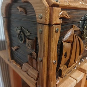 May include: Wooden treasure chest with carved nautical designs. The chest features a dark stained body with lighter wood accents, metal hardware, and a decorative lock. The chest is sitting on a wooden stand.