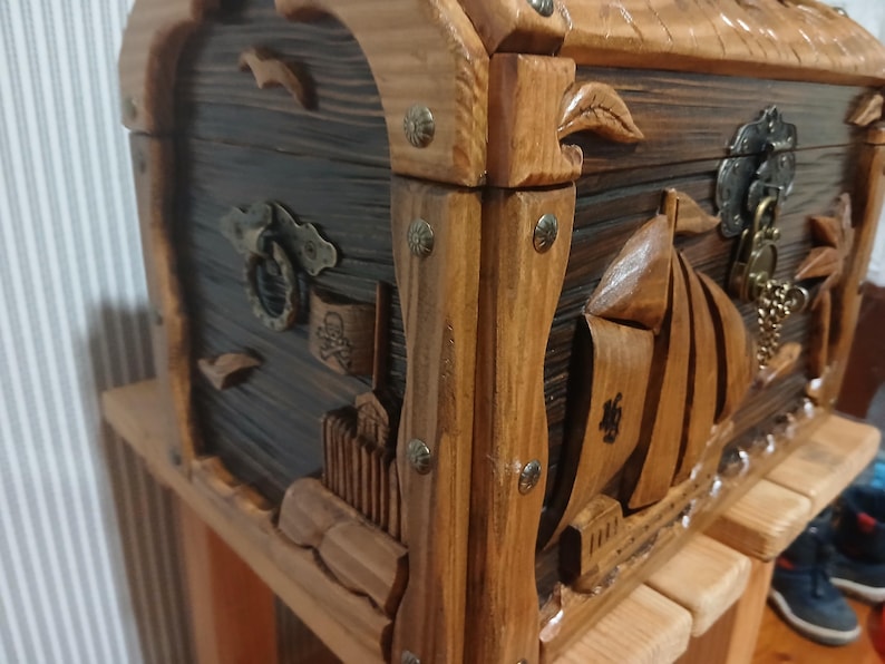 May include: Wooden treasure chest with carved nautical designs. The chest features a dark stained body with lighter wood accents, metal hardware, and a decorative lock. The chest is sitting on a wooden stand.