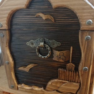 May include: Wooden treasure chest with a dark brown front and lighter brown frame. The chest features carved details including seagulls, a pirate flag, and a skull and crossbones. The chest has a metal clasp and decorative metal studs.