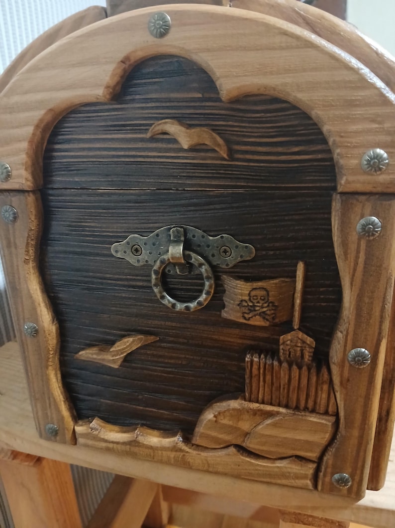 May include: Wooden treasure chest with a dark brown front and lighter brown frame. The chest features carved details including seagulls, a pirate flag, and a skull and crossbones. The chest has a metal clasp and decorative metal studs.