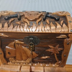 May include: Wooden treasure chest with carved pirate ship, palm tree, and skull designs. The chest has a dark brown finish with metal accents and a key. The chest is sitting on a wooden stand.