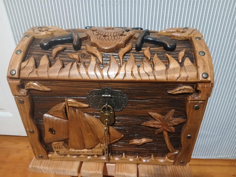 May include: Wooden treasure chest with carved pirate ship, palm tree, and skull designs. The chest has a dark brown finish with metal accents and a key. The chest is sitting on a wooden stand.