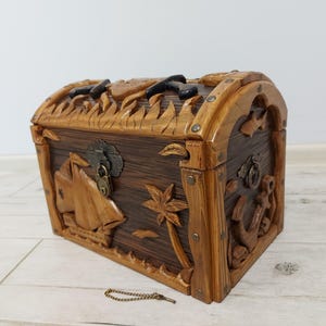 May include: A wooden treasure chest with a domed lid and intricate carvings. The chest features nautical-themed designs, including a sailboat, palm tree, and anchor. The chest is brown with lighter wood accents and metal hardware.