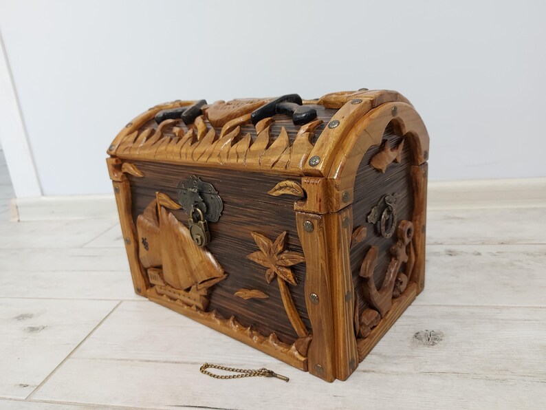 May include: A wooden treasure chest with a domed lid and intricate carvings. The chest features nautical-themed designs, including a sailboat, palm tree, and anchor. The chest is brown with lighter wood accents and metal hardware.