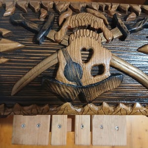 May include: Wooden chest with intricate carvings. The chest features a central design of a stylized skull with horns, axes, and other Viking-themed elements. The wood has a dark stain with lighter carved details.
