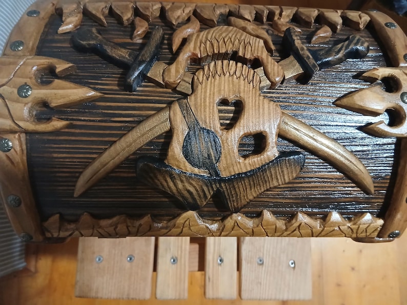 May include: Wooden chest with intricate carvings. The chest features a central design of a stylized skull with horns, axes, and other Viking-themed elements. The wood has a dark stain with lighter carved details.