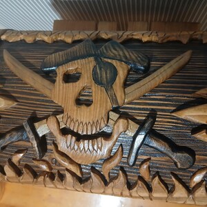 May include: Wooden pirate-themed chest with a carved skull and crossbones design. The chest features crossed swords, a pirate hat, and flames carved along the bottom. The chest is made of wood with a natural finish and dark accents.