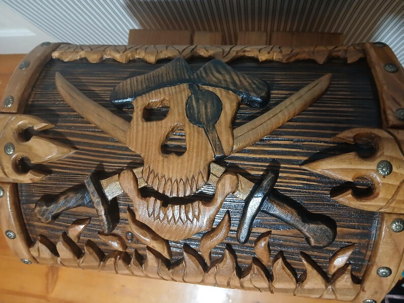 May include: Wooden pirate-themed chest with a carved skull and crossbones design. The chest features crossed swords, a pirate hat, and flames carved along the bottom. The chest is made of wood with a natural finish and dark accents.