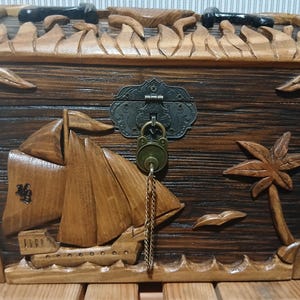 May include: Wooden treasure chest with carved sailboat, palm tree, and decorative lock. The chest is made of dark and light brown wood, with metal accents. The chest is approximately 12 inches wide and 8 inches tall.