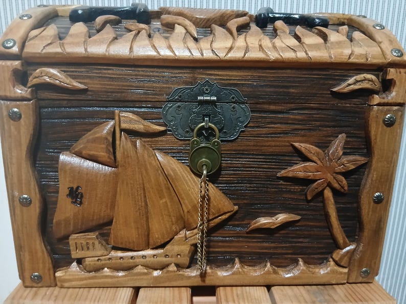 May include: Wooden treasure chest with carved sailboat, palm tree, and decorative lock. The chest is made of dark and light brown wood, with metal accents. The chest is approximately 12 inches wide and 8 inches tall.