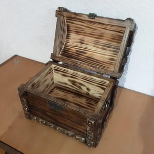 May include: A wooden box with a hinged lid, the box is made of dark wood with a burnt finish and has intricate carvings around the edges.