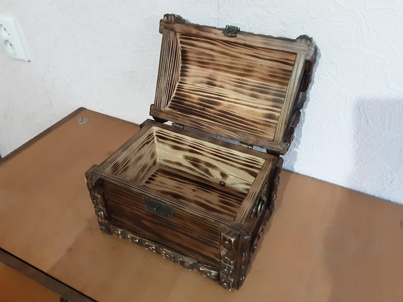 May include: A wooden box with a hinged lid, the box is made of dark wood with a burnt finish and has intricate carvings around the edges.