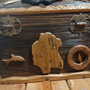 May include: Wooden treasure chest with a dark stained finish and lighter wood trim. Features include metal hinges and clasps, a carved map, a compass, and a dolphin. The chest is designed with a rustic, nautical theme.