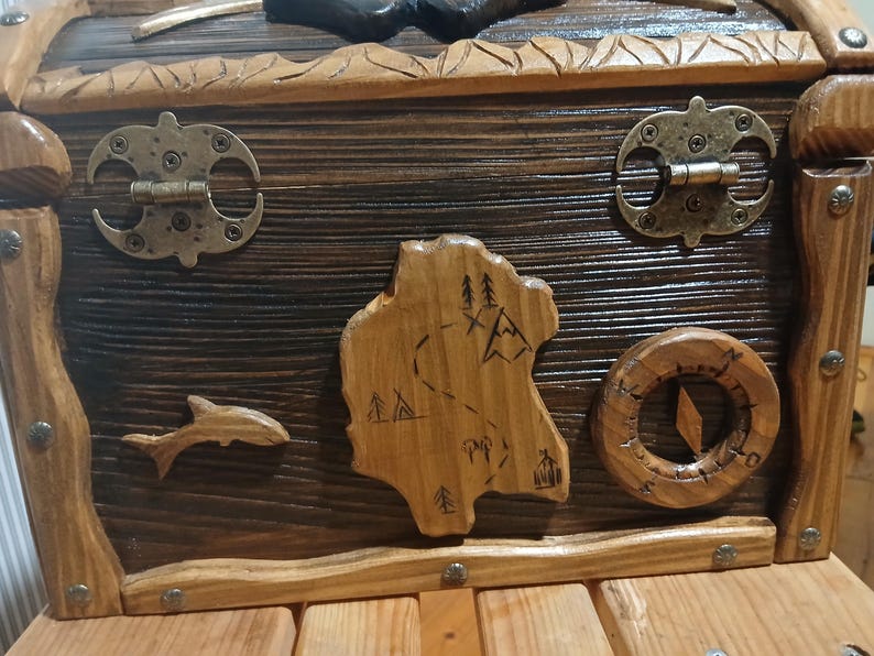 May include: Wooden treasure chest with a dark stained finish and lighter wood trim. Features include metal hinges and clasps, a carved map, a compass, and a dolphin. The chest is designed with a rustic, nautical theme.