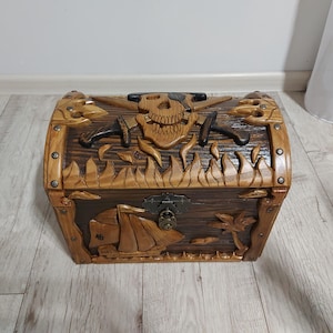 May include: A wooden treasure chest with intricate carvings. The chest features a skull and crossed swords design on the front, with a ship and palm tree carved on the lower section. The chest is brown with a dark brown interior.