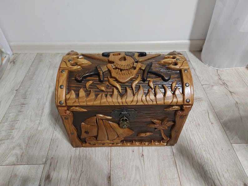 May include: A wooden treasure chest with intricate carvings. The chest features a skull and crossed swords design on the front, with a ship and palm tree carved on the lower section. The chest is brown with a dark brown interior.