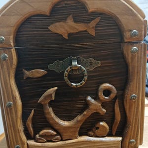 May include: Wooden treasure chest with carved nautical elements. Features a shark, fish, anchor, and skull design. The chest has a dark brown interior and a lighter brown frame, with metal accents. A decorative storage box.