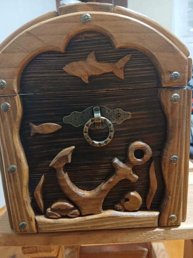 May include: Wooden treasure chest with carved nautical elements. Features a shark, fish, anchor, and skull design. The chest has a dark brown interior and a lighter brown frame, with metal accents. A decorative storage box.