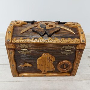 May include: A wooden treasure chest with a dark brown finish and carved details. The chest features a map, compass, and fish design on the front. The lid has a carved skull and crossed swords. The chest is 12 inches long, 8 inches wide, and 8 inches tall.